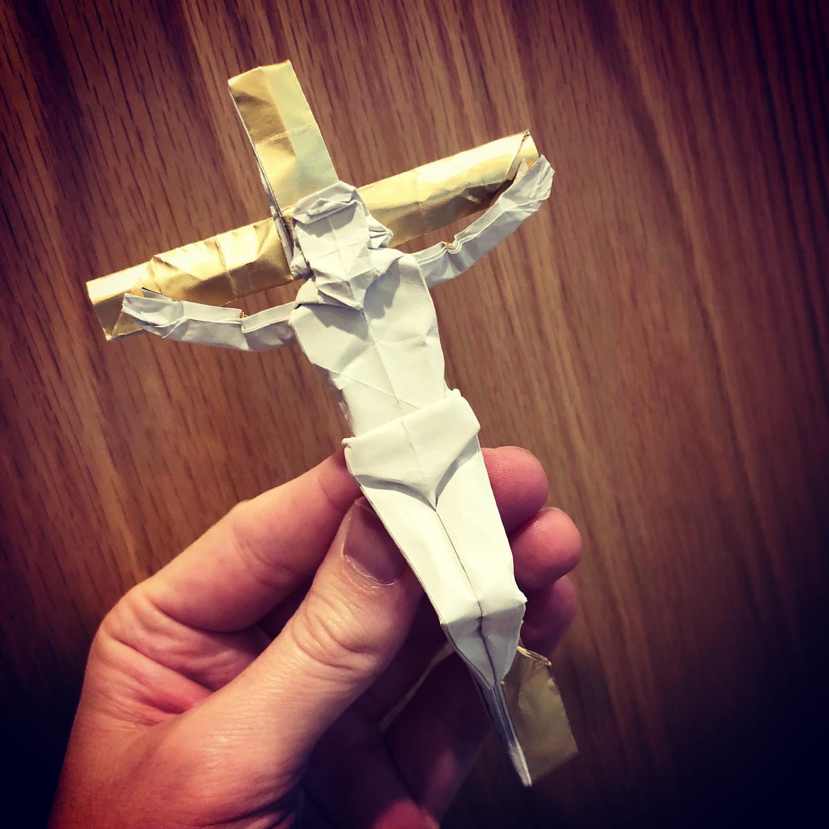 An origami crucifix being held in a hand over a wooden background. Designed and folded by Matthew Green.