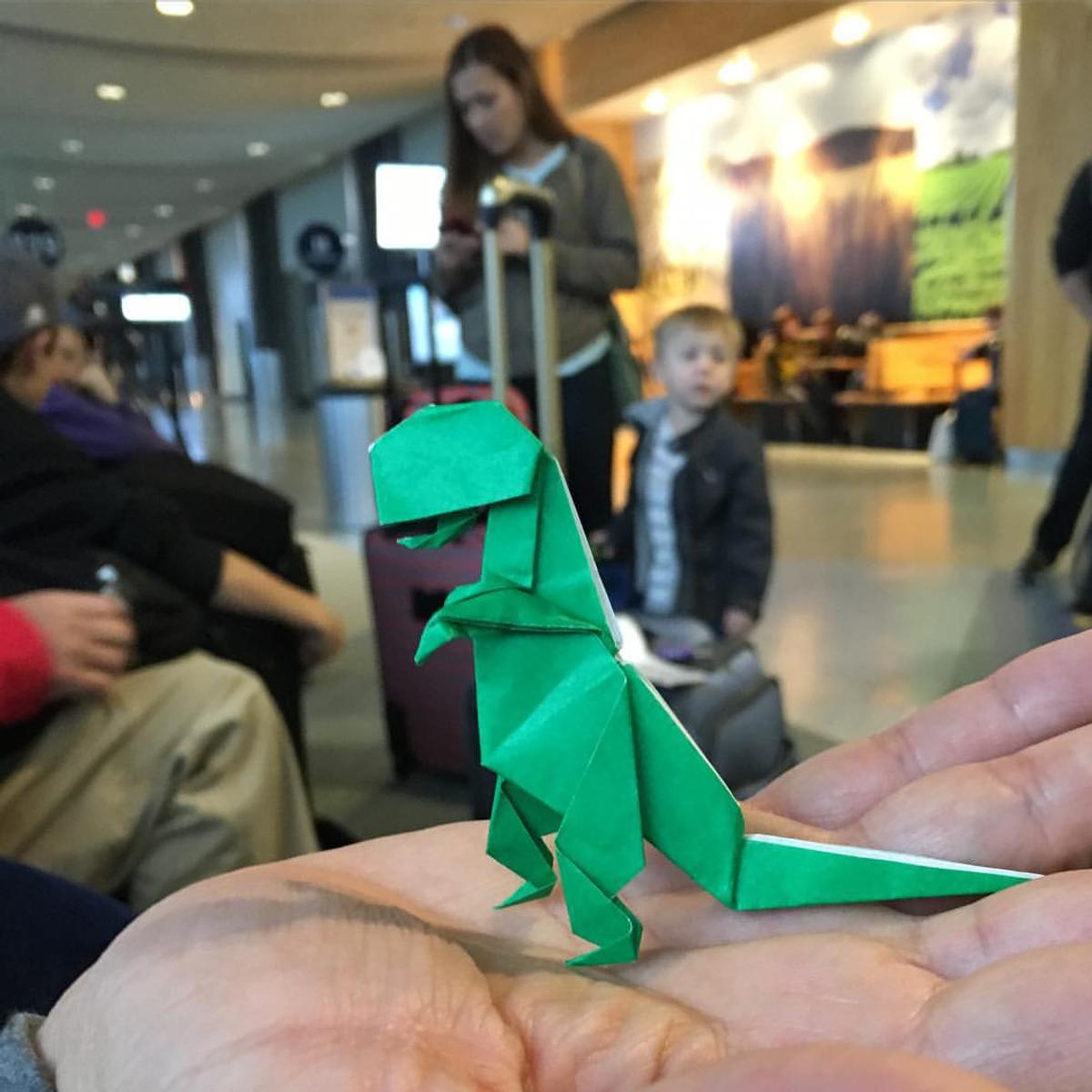 An origami Tyrannosaurus Rex, held in the palm of a hand, with passengers blurrily visible int he background. The photo is taken in an airport.