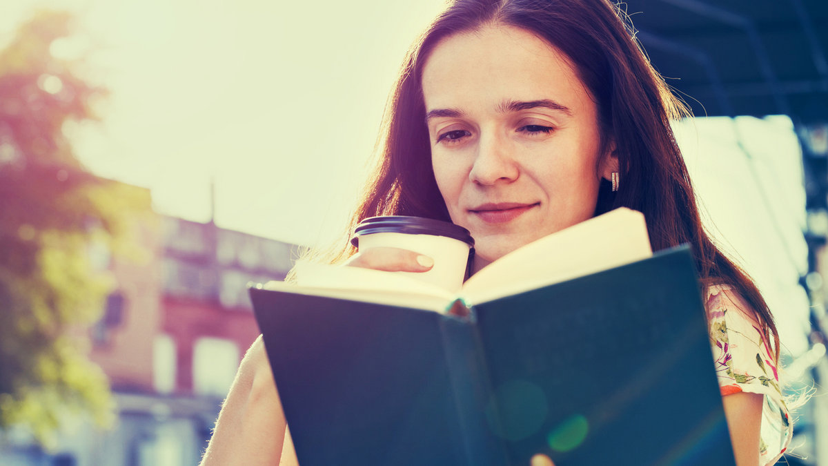 Smiling girl sitting at summer terrace in street with morning coffee and reading book
