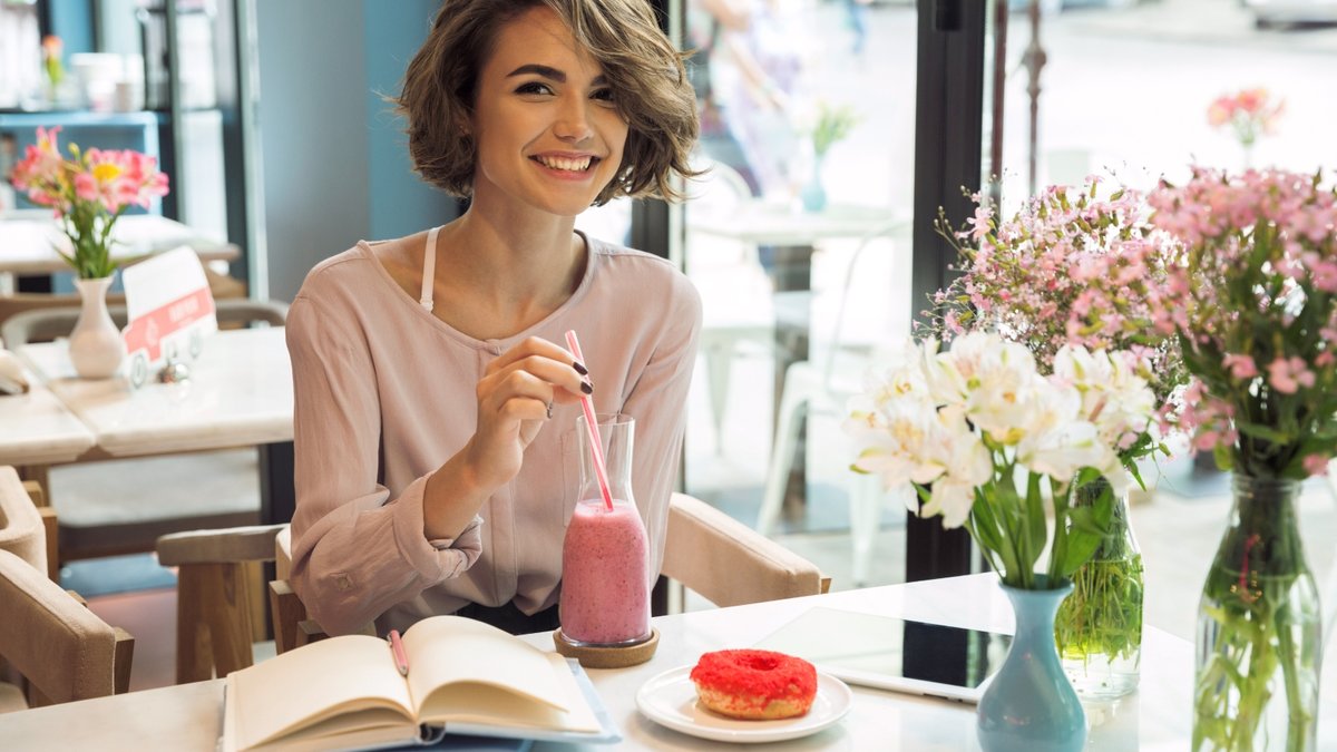 uśmiechnięta kobieta siedzi w kawiarni i pije smoothie, a przed nią leżą książki i pączek na talerzu
