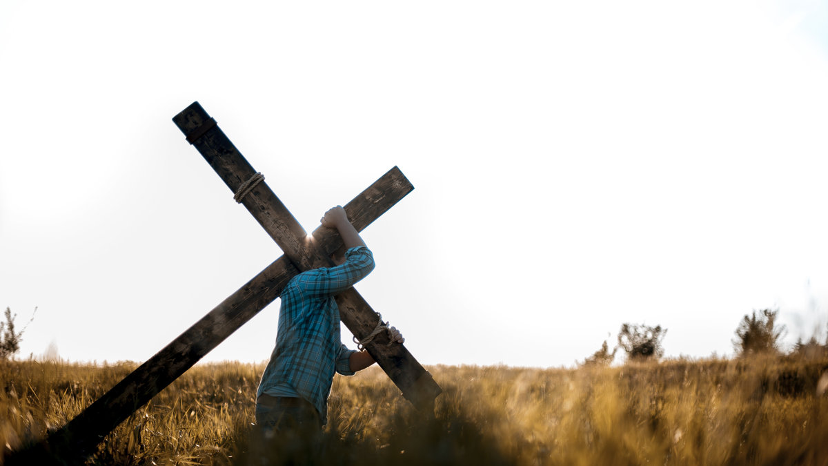 A-male-carrying-a-handmade-wooden-cross-on-his-back-in-a-grassy-field-shutterstock_1553325437.jpg