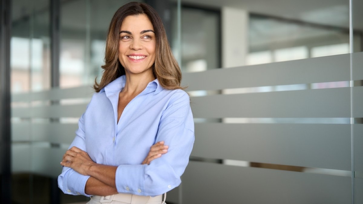 Happy confident mature professional business woman standing at work in office arms crossed looking away