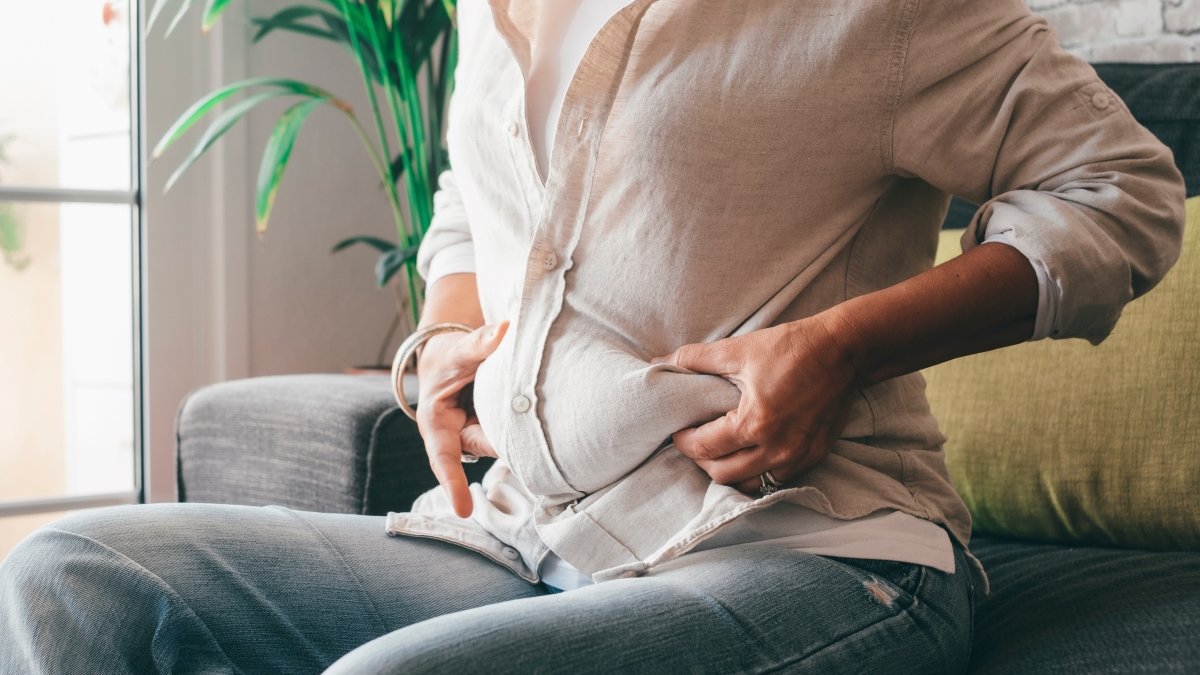 Midsection of woman hands holding her belly fat sitting on sofa at home.