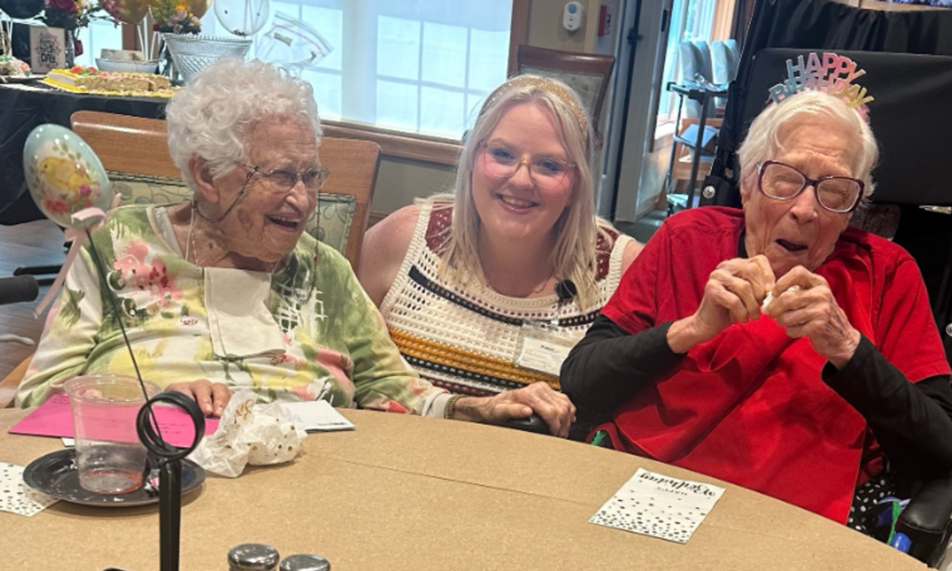 two elderly women with a blonde younger woman in the middle. one has a happy birthday hat