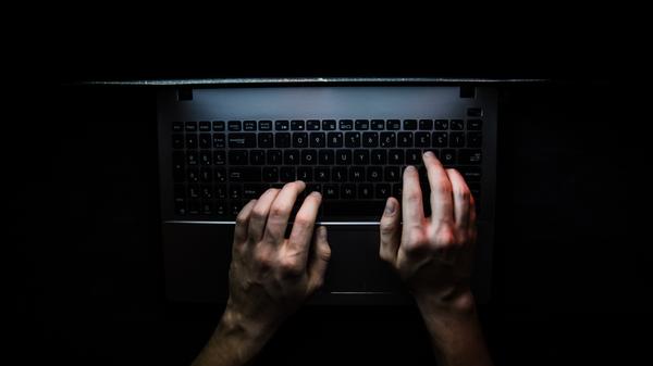 Dłonie internauty widziane z góry oparte na klawiaturze laptopa w zaciemnionym pokoju