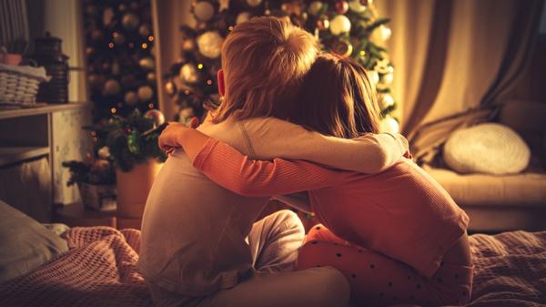 Children sit hugging in bed and look at the Christmas tree