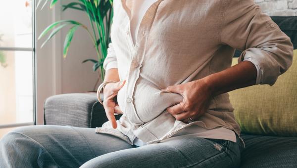 Midsection of woman hands holding her belly fat sitting on sofa at home.