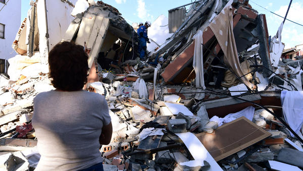 A woman looks at rescuers searching for survivors amid the rubble of a collapsed building after an earthquake in Pereira, Colombia on August 10, 2026. A magnitude 7.4 earthquake struck Colombia on August 10, 2026, and killed at least 111 people, damaged more than 1,500 buildings, and left an unknown number of people buried under rubble. The government declared a state of emergency to attend to the quake that shook several cities across the coffee-producing region, the Pacific coast, and neighboring Latin American countries, sending terrified residents racing into the street.