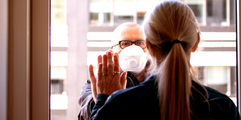 web3-a-married-couple-separated-by-corona-covid-19-virus.-she-is-quarantined-indoors-and-he-is-outside-wearing-protective-mask.-they-are-holding-hands-through-a-window-glass-looking-lovi.jpg